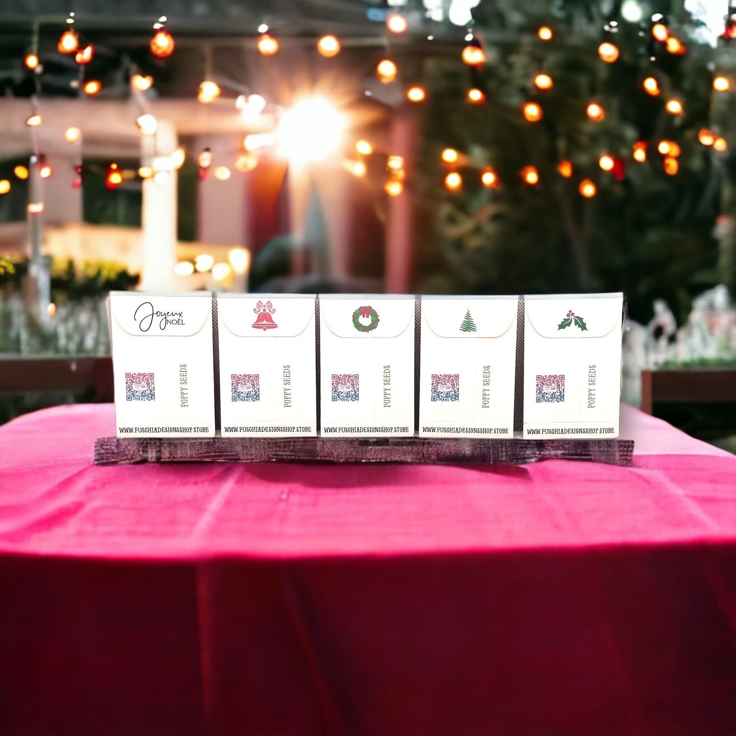 Backs of five French Christmas poppy seed packets in a row on a red tablecloth outdoors, with bell, wreath, tree and holly designs on the flaps, QR codes and the Fuschia Designs Shop website