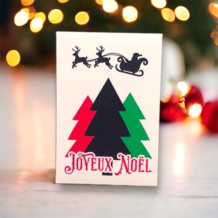French Christmas seed packet with a silhouette of Santa's sleigh above three stylized Christmas trees in red, black and green, reading 'Joyeux Noël', in front of Christmas tree lights