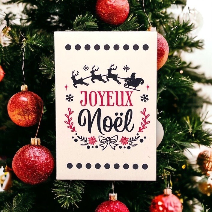 French Christmas seed packet reading 'Joyeux Noël' with a silhouette of three reindeer pulling Santa's sleigh, propped in a Christmas tree among red glitter baubles