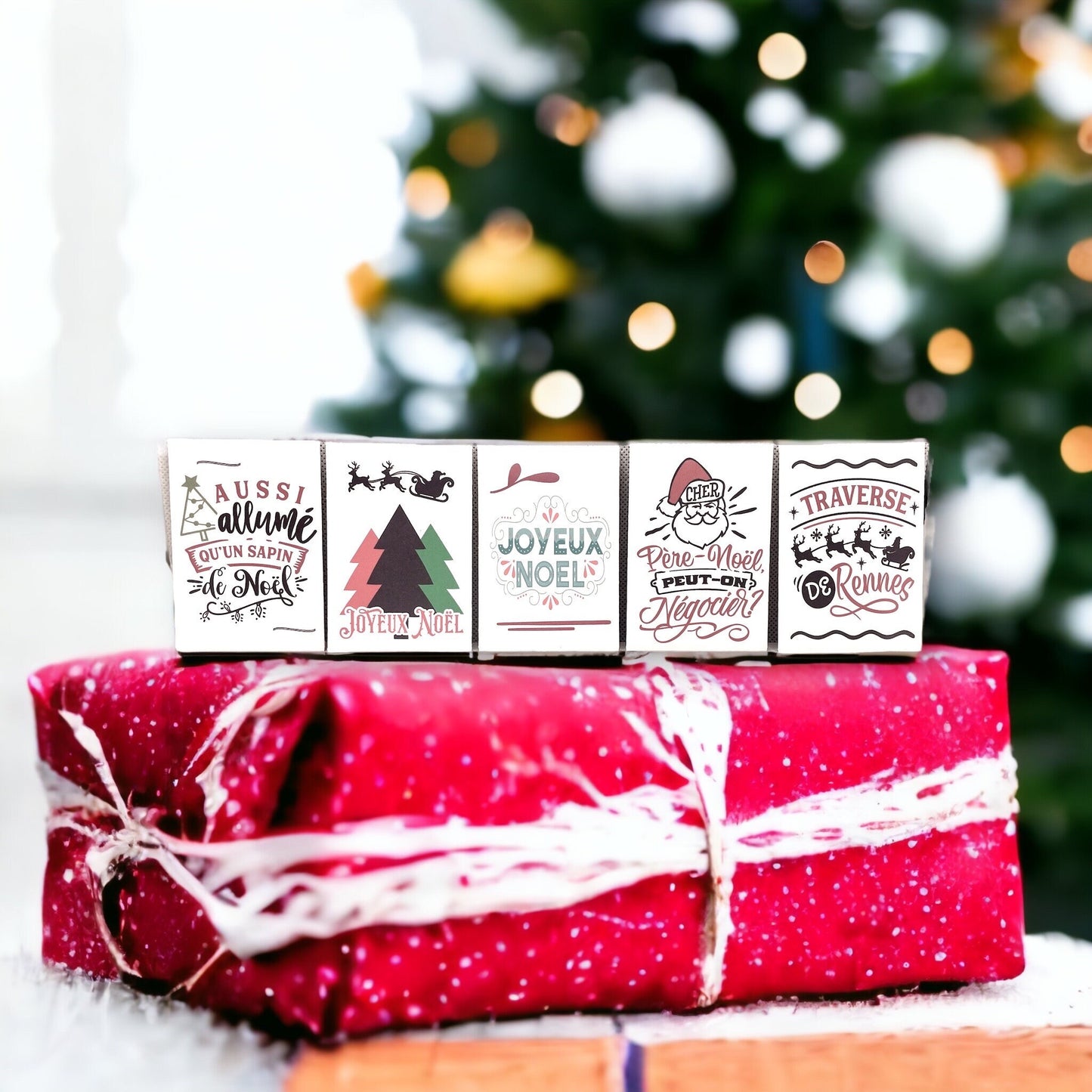 Five French Christmas seed packets standing on a red wrapped gift in front of a Christmas tree, reading 'Aussi allumé qu'un sapin de Noël', 'Joyeux Noël', 'Cher Père-Noël, peut-on négocier?' and 'Traverse de Rennes'