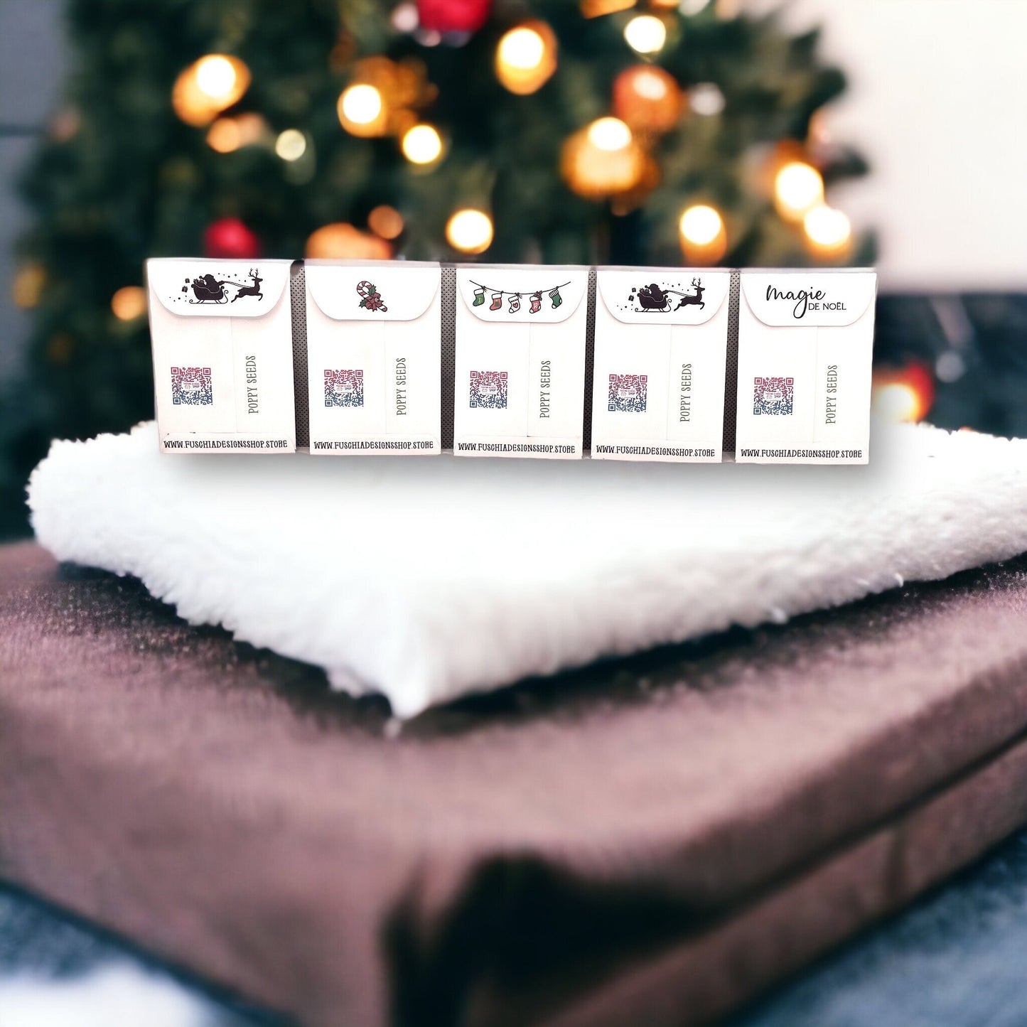 Backs of five French Christmas poppy seed packets in a row on a white throw, showing Santa's sleigh, candy cane, stocking and 'Magie de Noël' designs on the flaps, QR codes and the Fuschia Designs Shop website