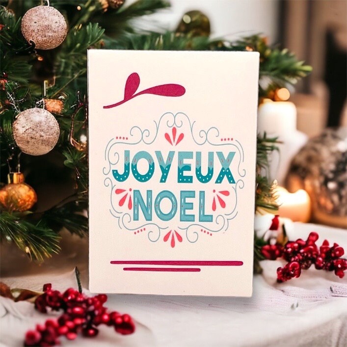 French Christmas seed packet reading 'Joyeux Noël' in teal lettering with pink flourishes, in front of a Christmas tree with baubles, berries and candles