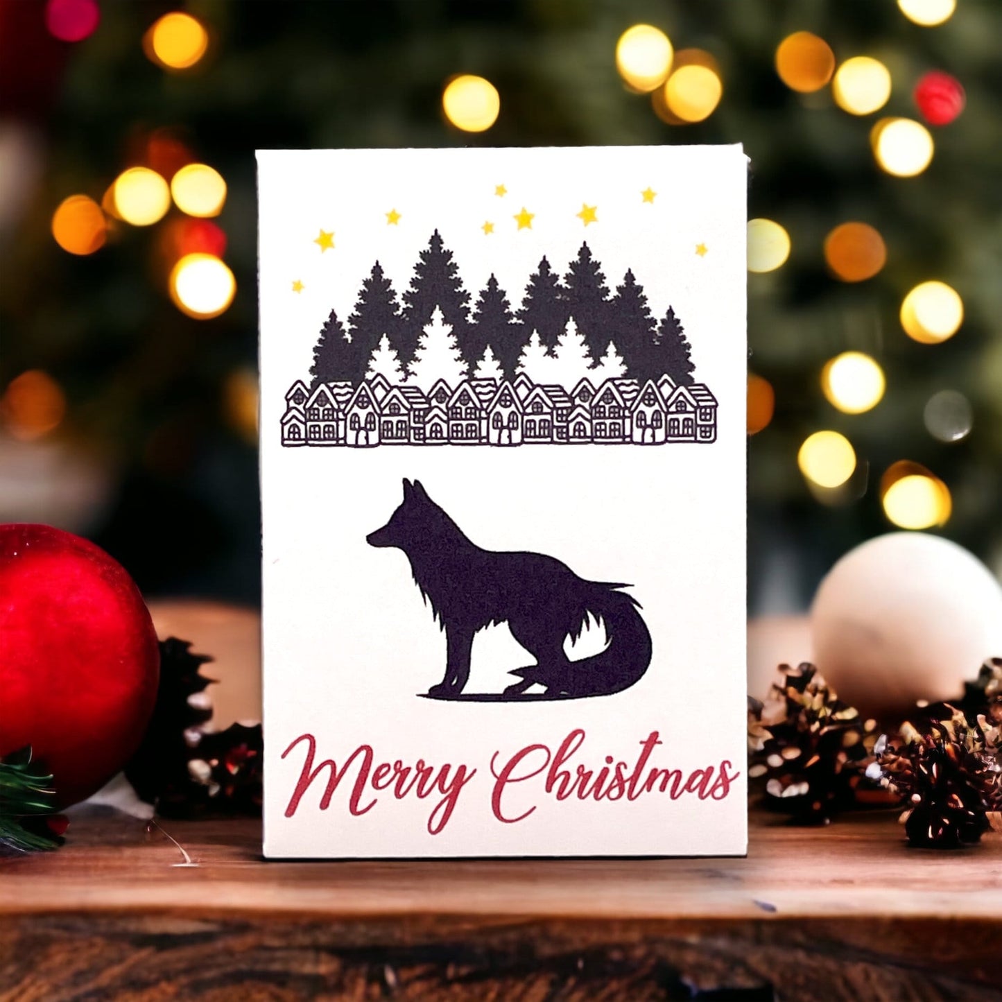 Woodland Christmas seed packet showing a row of village houses under pine trees and stars, above a black fox silhouette and the words 'Merry Christmas', beside baubles and pine cones