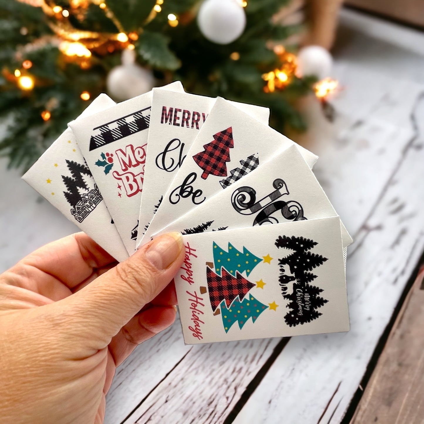 Hand holding a fanned set of woodland Christmas seed packets in front of a Christmas tree, the front one showing plaid and teal Christmas trees and deer among pine trees with the words 'Happy Holidays'