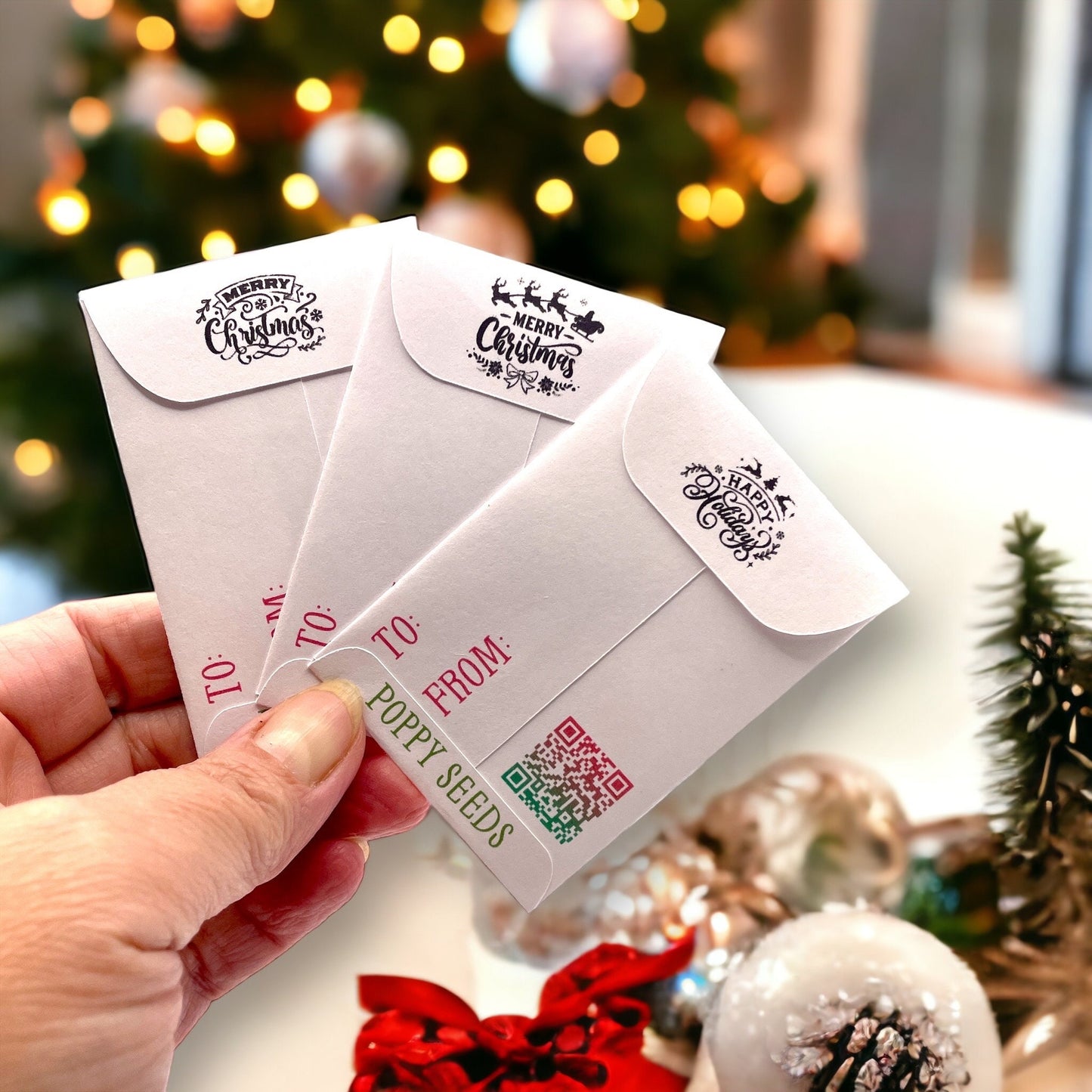 Hand holding the backs of three vintage Christmas seed packets, with 'Merry Christmas' and 'Happy Holidays' stamps on the flaps, To and From lines, a QR code and 'Poppy Seeds'