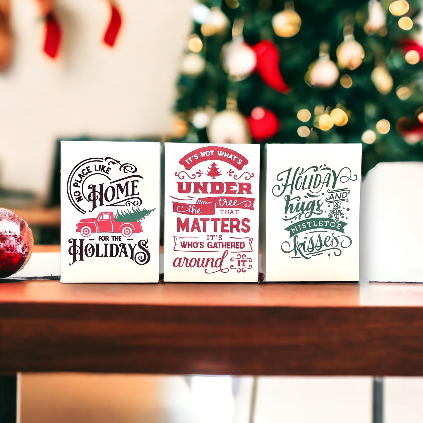 Three vintage-style Christmas seed packets on a wooden ledge in front of a Christmas tree, reading 'No place like Home for the Holidays', 'It's not what's under the tree that matters, it's who's gathered around it' and 'Holiday hugs and mistletoe kisses'