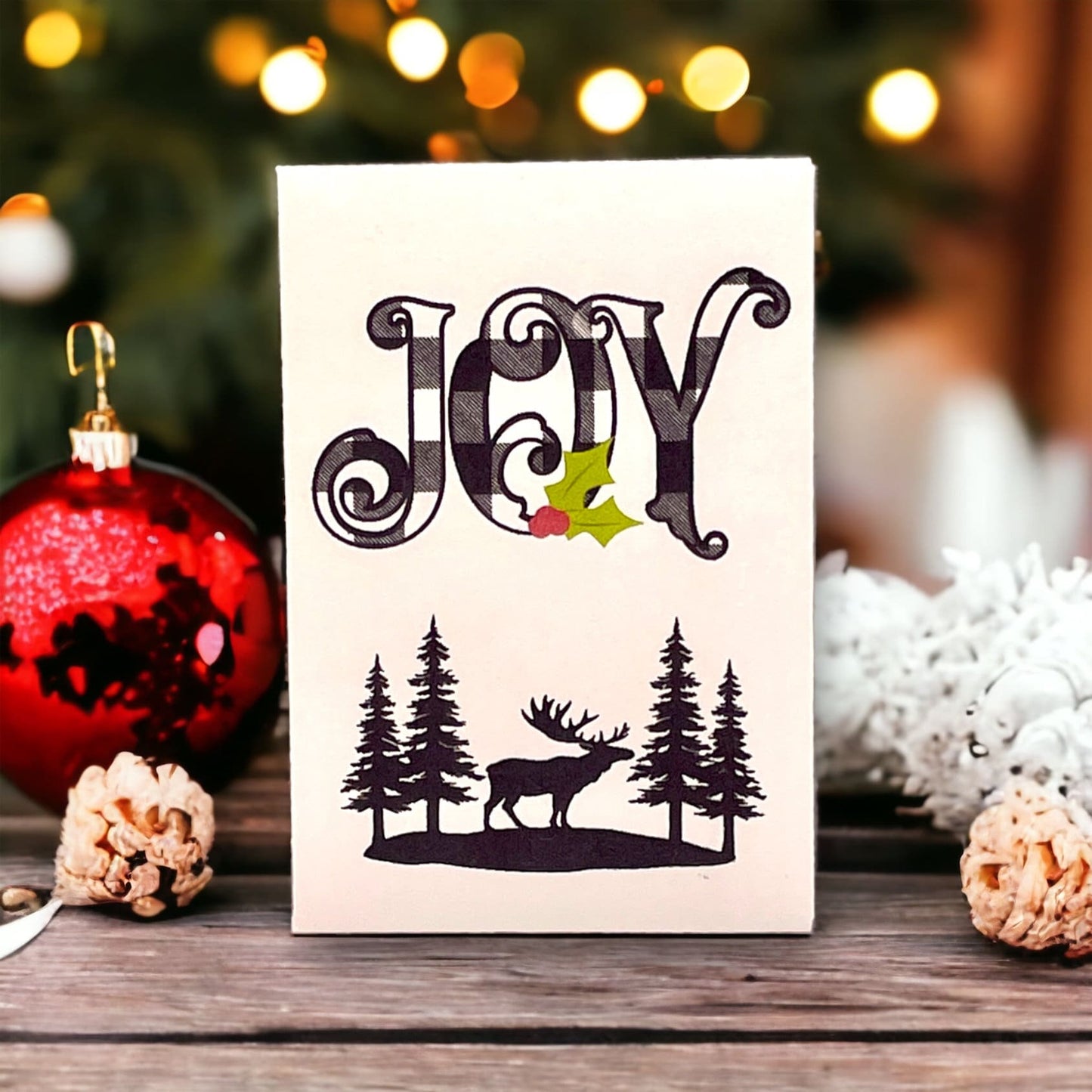 Woodland Christmas seed packet with the word 'JOY' in black buffalo plaid above a moose silhouette standing between pine trees, beside a red bauble and pine cones