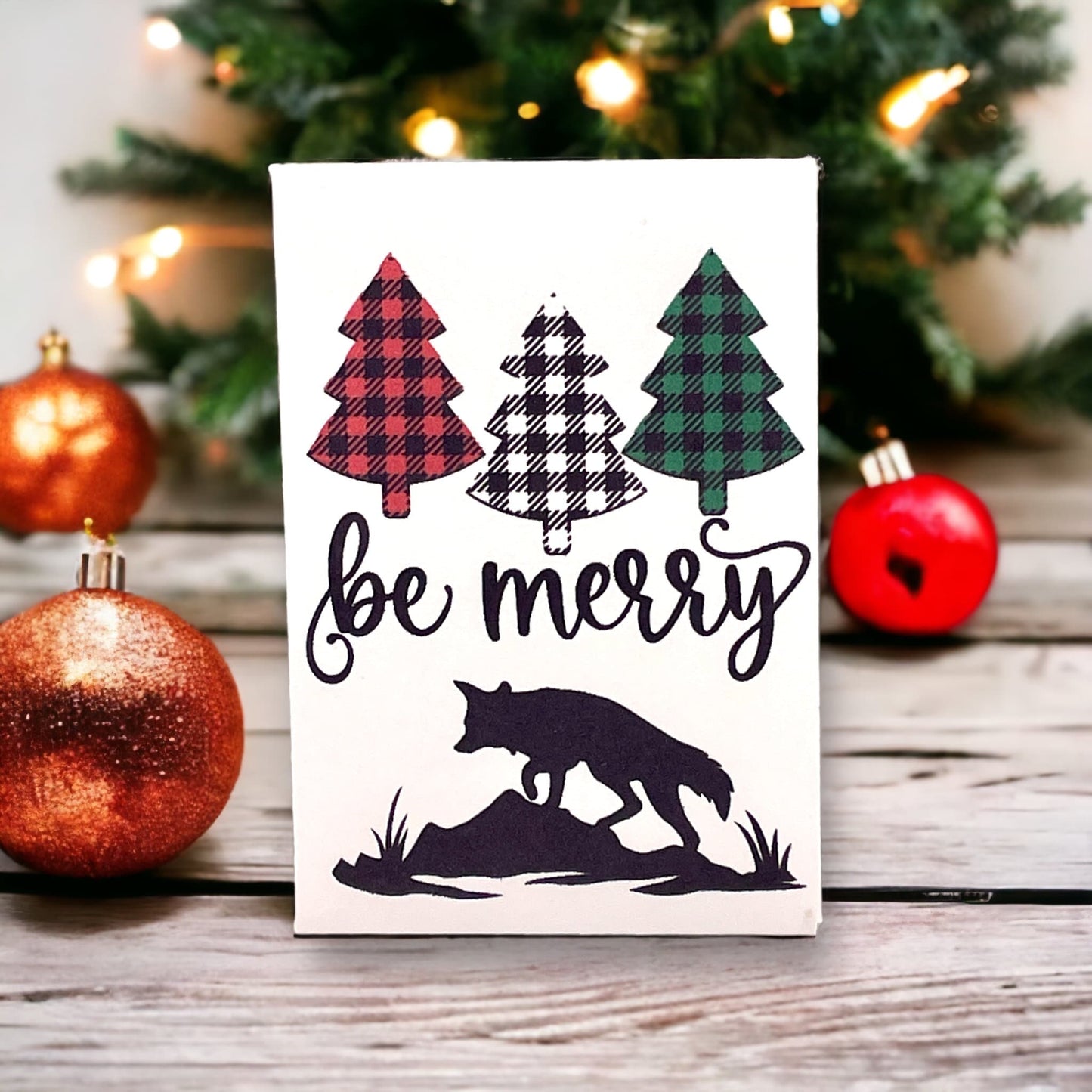 Woodland Christmas seed packet with three buffalo-plaid pine trees in red, black and green above the words 'be merry' and a fox silhouette, beside copper and red baubles