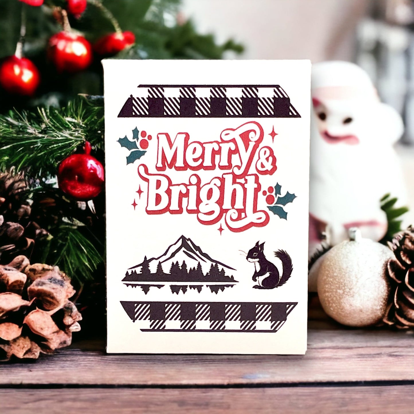 Woodland Christmas seed packet with buffalo-plaid borders reading 'Merry & Bright', with a mountain, forest and lake silhouette and a squirrel, beside pine cones and a bauble