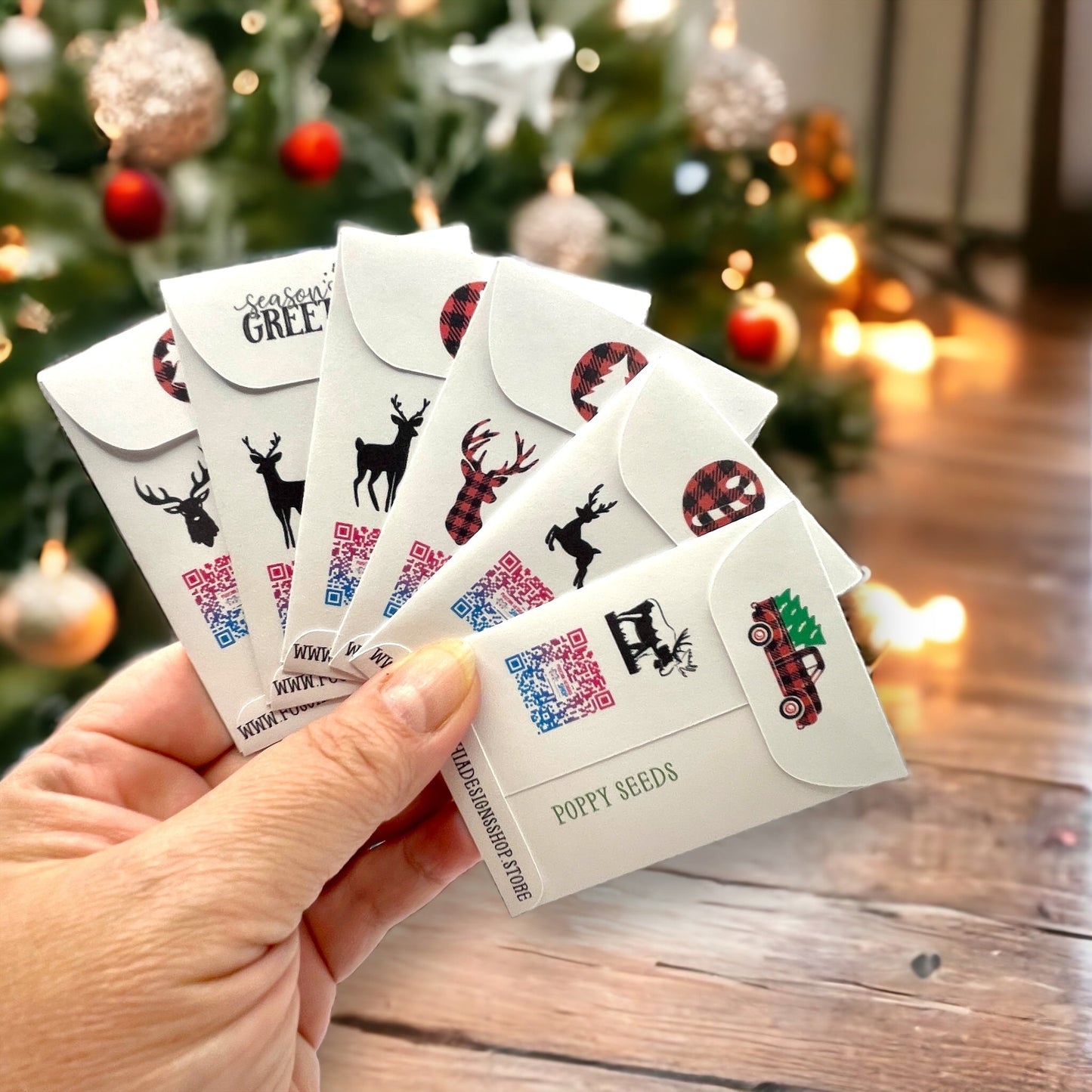 Hand holding the backs of woodland Christmas seed packets in front of a Christmas tree, showing deer and moose silhouettes, QR codes, buffalo-plaid flap designs and 'Poppy Seeds'