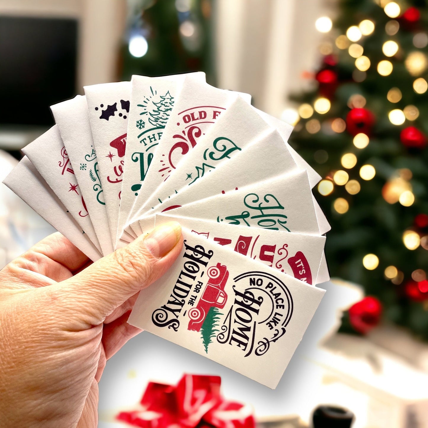 Hand holding a fanned set of vintage-style Christmas seed packets in front of a lit Christmas tree, the front one showing a red truck carrying a tree with the words 'No place like Home for the Holidays'