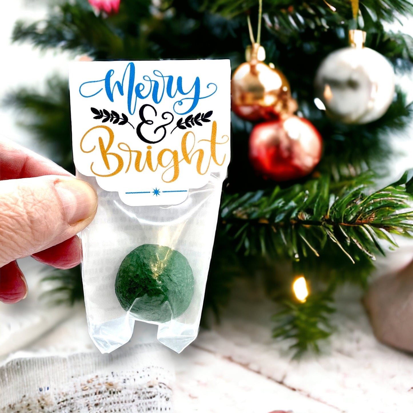 Hand holding an individually wrapped green Christmas seed bomb with a 'Merry & Bright' card topper, in front of Christmas baubles
