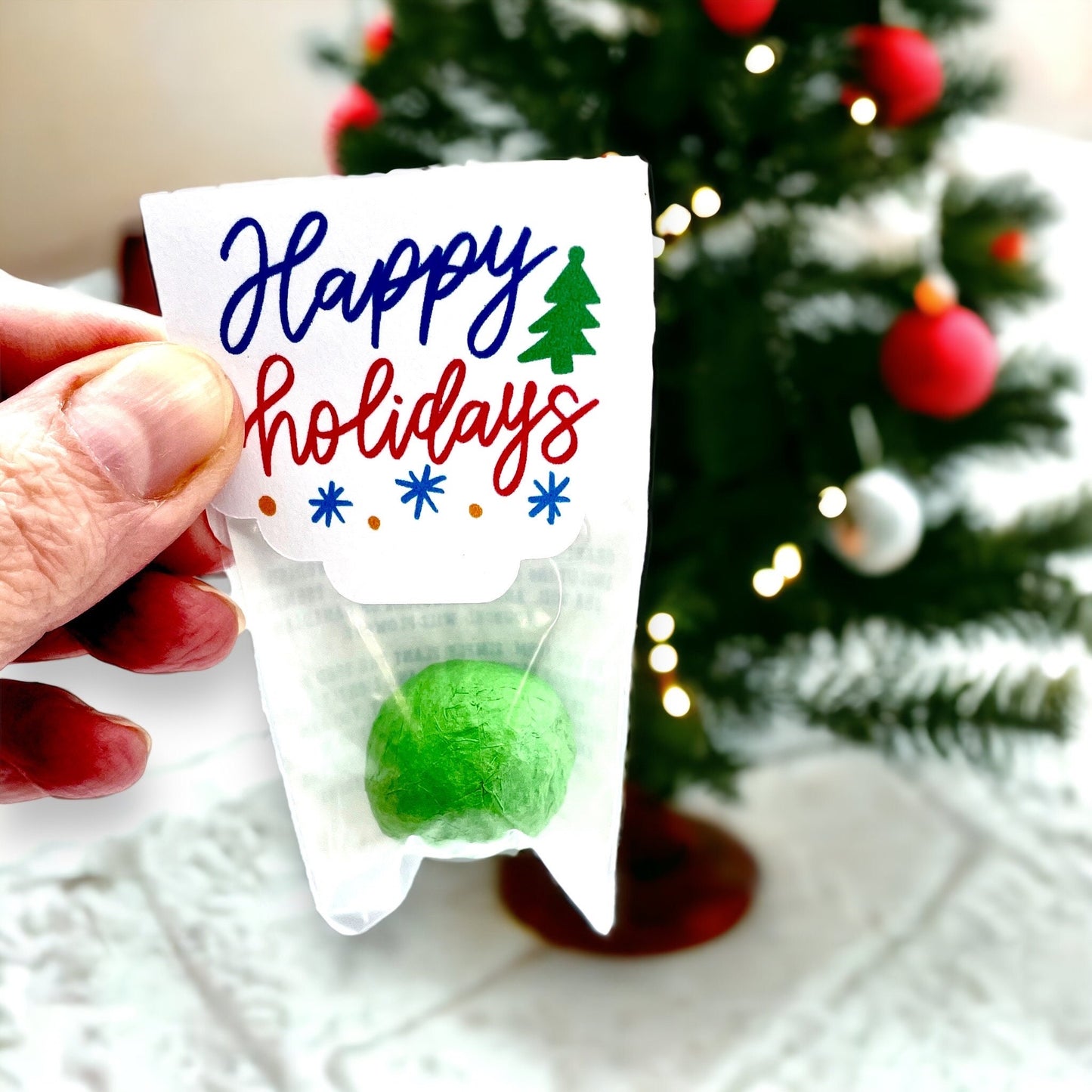 Hand holding an individually wrapped green Christmas seed bomb with a 'Happy Holidays' card topper in blue and red script, in front of a small Christmas tree