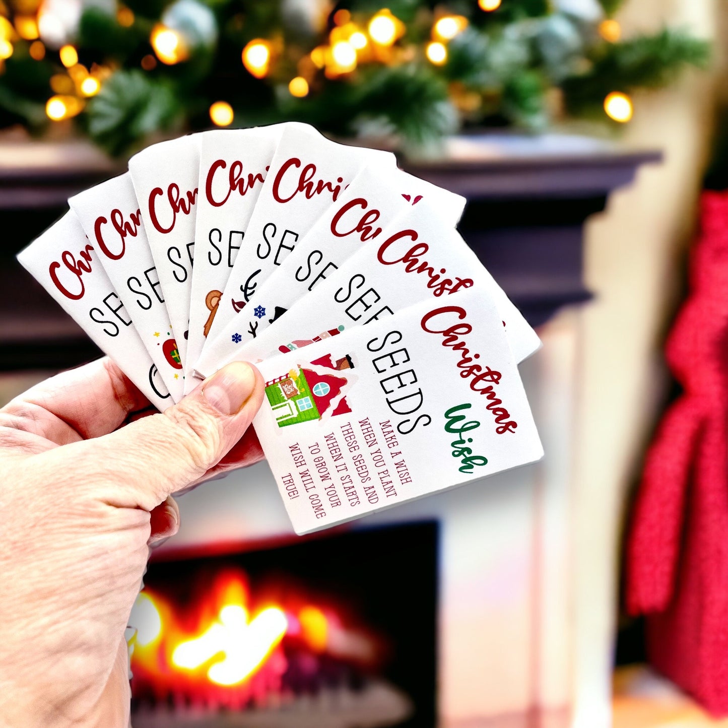 Hand holding a fanned set of 'Christmas Wish Seeds' packets in front of a fireplace and Christmas tree, the front packet showing a Santa's house design