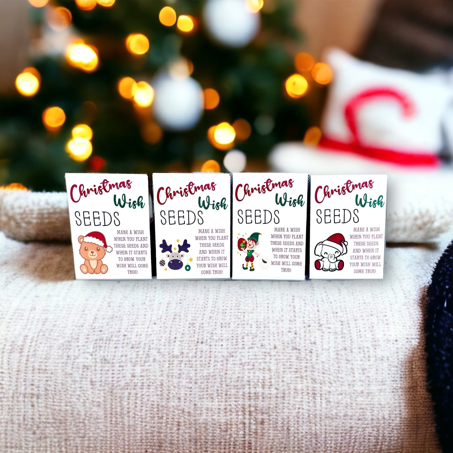 Four 'Christmas Wish Seeds' packets with teddy bear, moose, elf and elephant designs, arranged on a cushion in front of Christmas tree lights