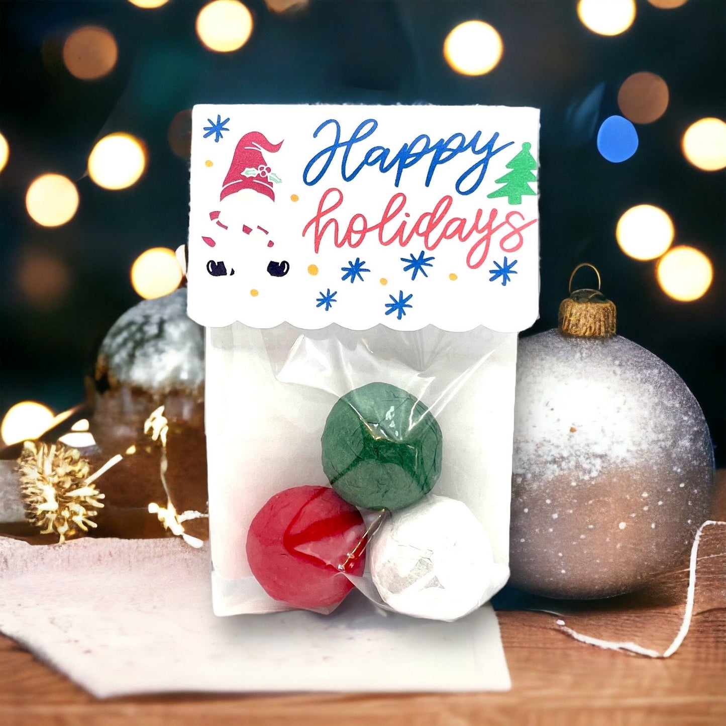 Clear bag of three Christmas seed bombs in green, red and white, with a 'Happy holidays' card topper decorated with a gnome and a Christmas tree, beside a frosted bauble