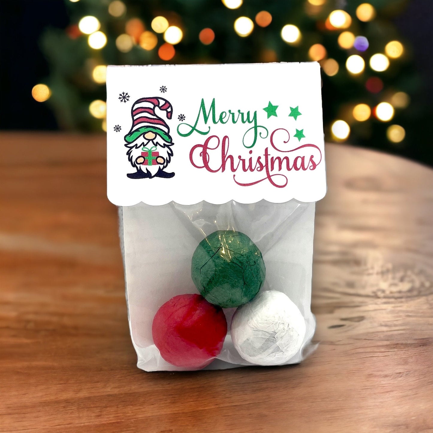 Clear bag of three Christmas seed bombs in green, red and white, with a 'Merry Christmas' card topper showing a gnome holding a gift, on a wooden table