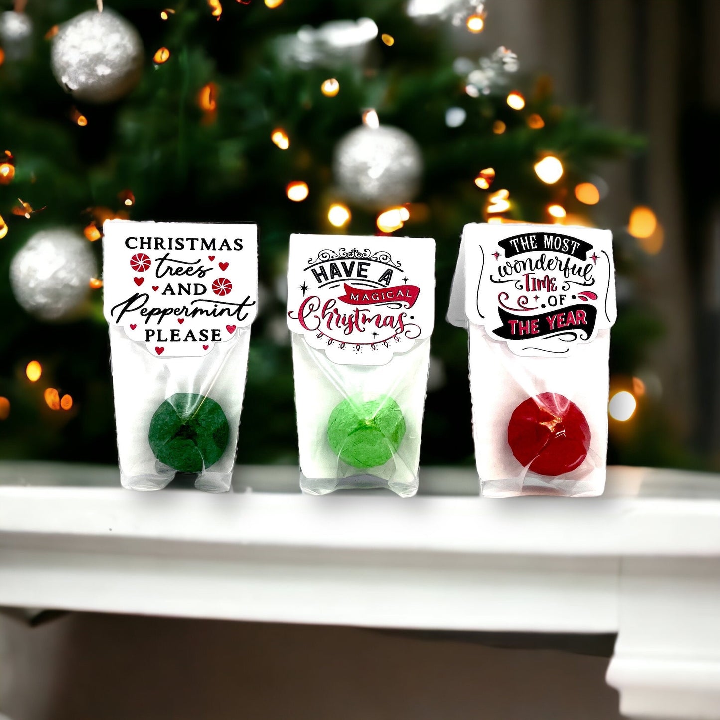 Three individually wrapped Christmas seed bombs on a white ledge in front of a lit Christmas tree, with toppers reading 'Christmas trees and Peppermint please', 'Have a Magical Christmas' and 'The most wonderful time of the year'