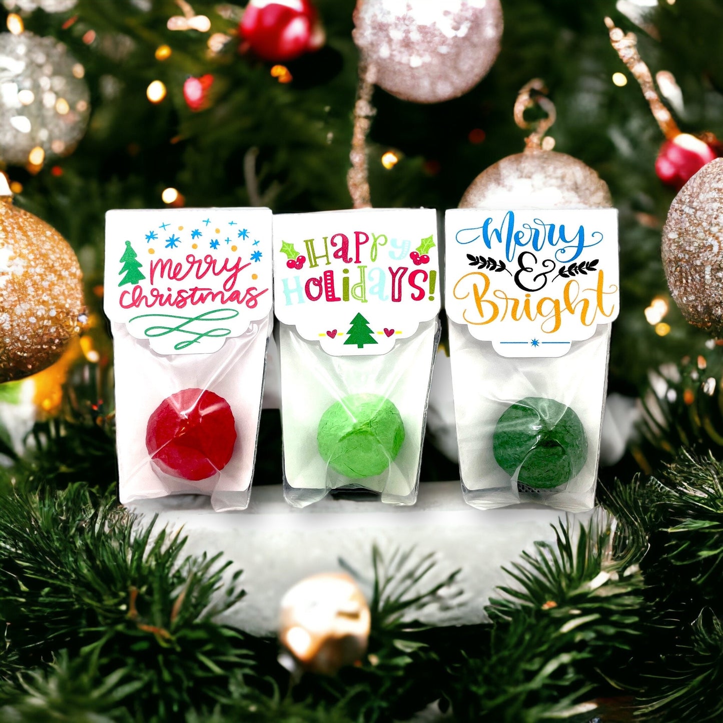 Three individually wrapped Christmas seed bombs in front of a decorated Christmas tree, with card toppers reading 'Merry Christmas', 'Happy Holidays!' and 'Merry & Bright'