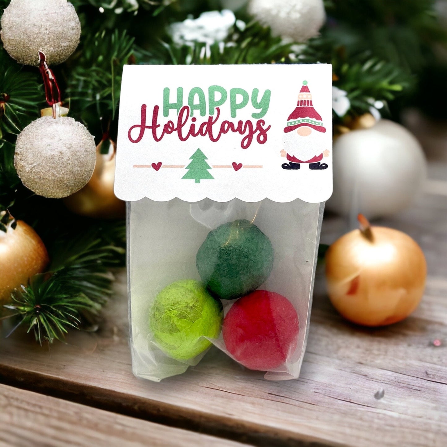 Clear bag of three Christmas seed bombs in green and red, with a 'Happy Holidays' card topper showing a small gnome, in front of a Christmas tree with baubles