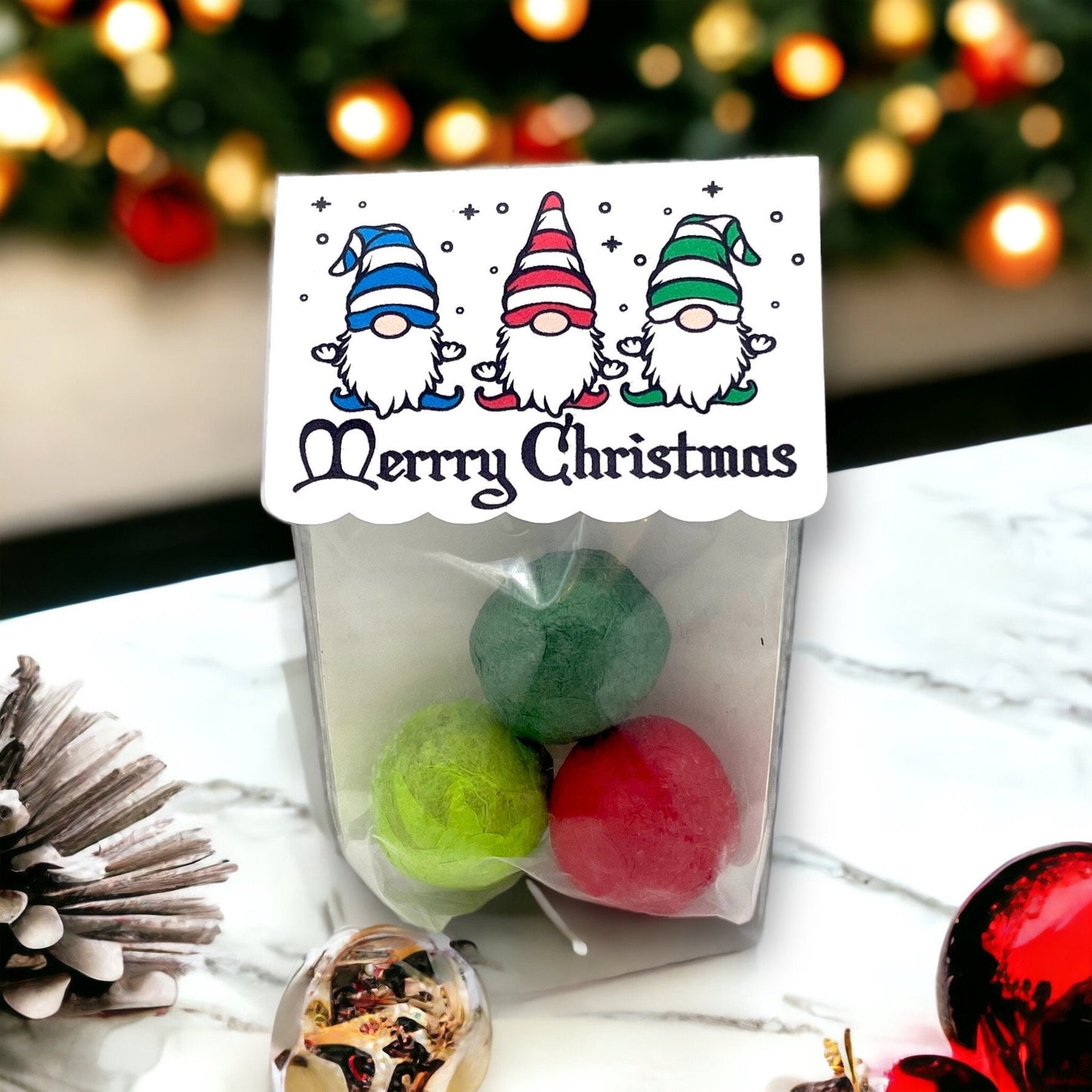 Clear bag of three Christmas seed bombs in green and red, with a card topper showing three gnomes in striped hats above the words 'Merrry Christmas', beside a pine cone and bauble