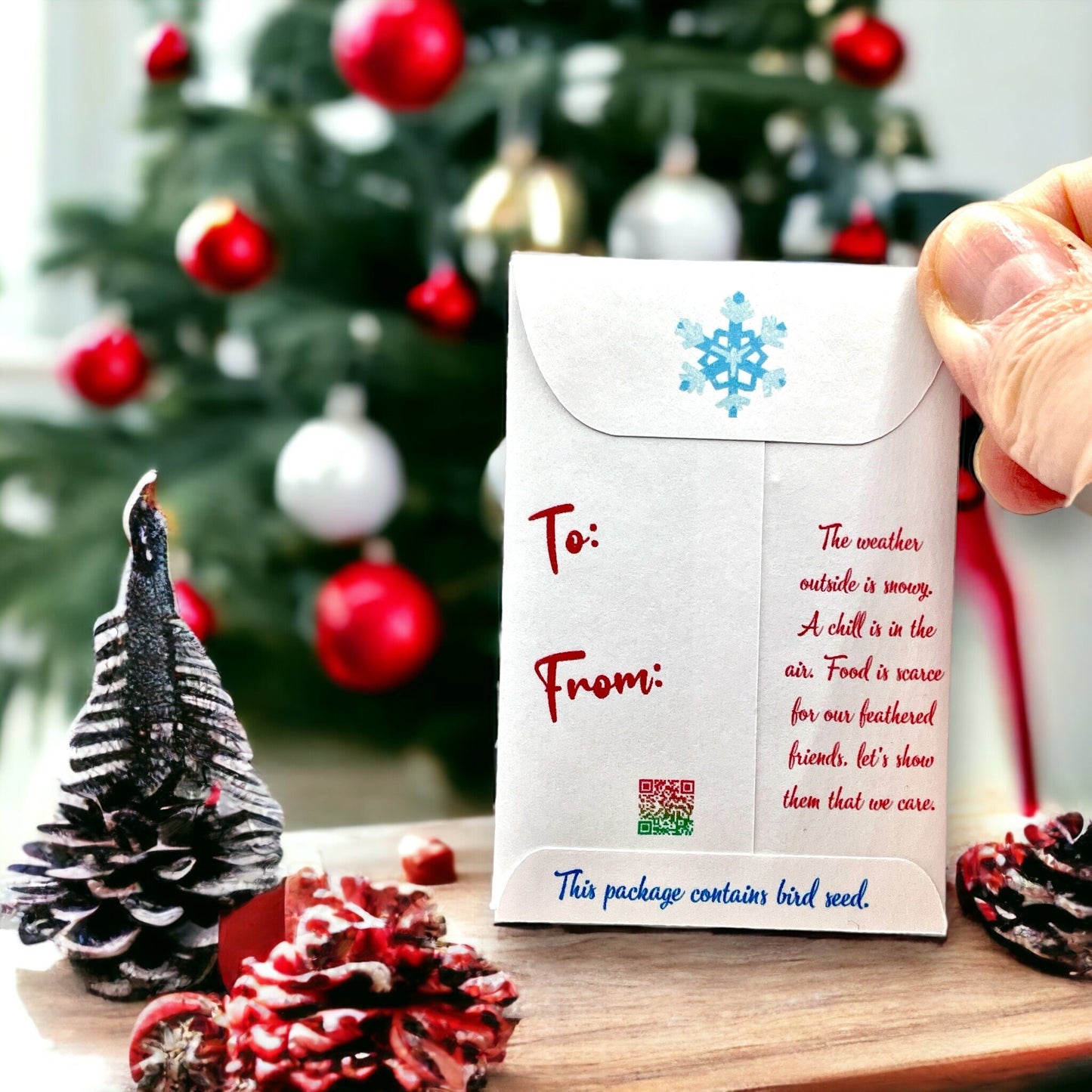 Hand holding the back of a Christmas bird seed packet in front of a Christmas tree, with To and From lines, a snowflake on the flap and a poem about feeding feathered friends in winter, ending 'This package contains bird seed'