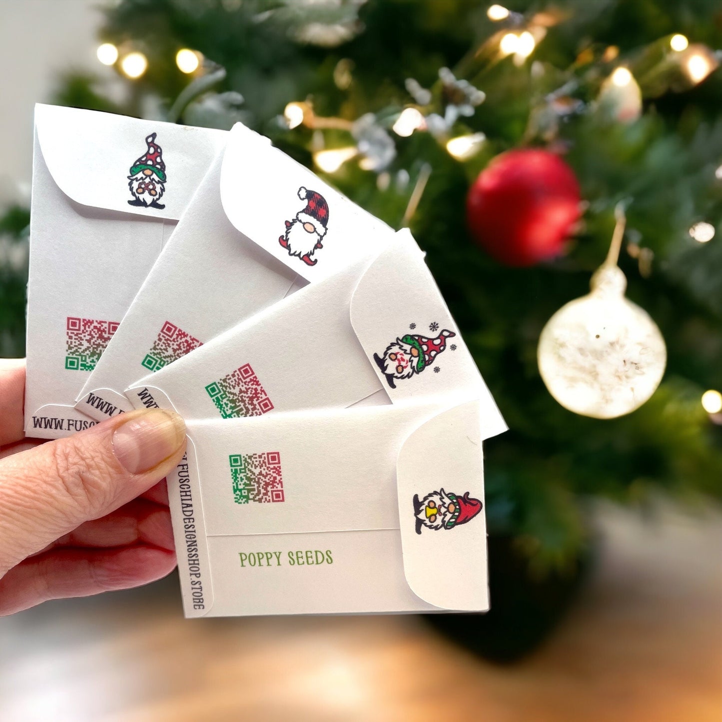 Hand holding the backs of Christmas gnome seed packets in front of a Christmas tree, showing small gnome illustrations on the flaps, QR codes, 'Poppy Seeds' and the Fuschia Designs Shop website