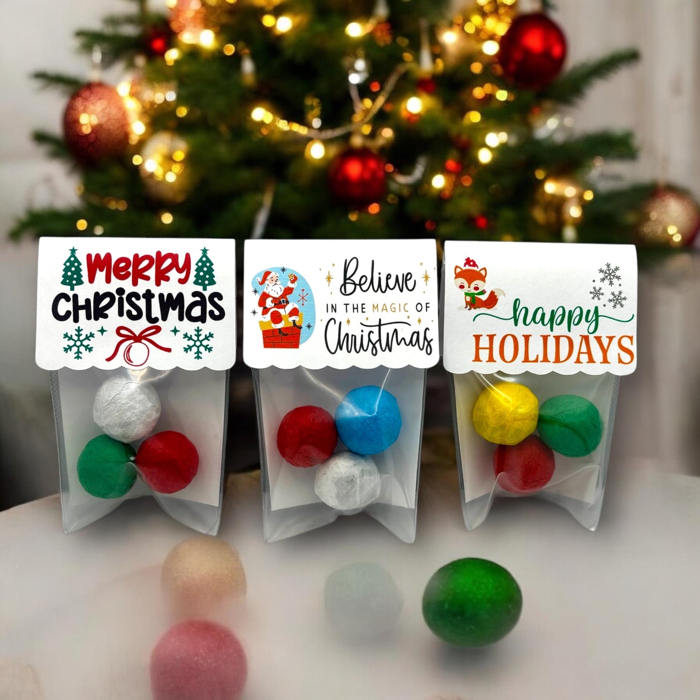 Three Christmas seed bomb 3-packs in front of a lit Christmas tree, with card toppers reading 'Merry Christmas', 'Believe in the magic of Christmas' with Santa, and 'happy HOLIDAYS' with a fox, each holding three colourful seed bombs
