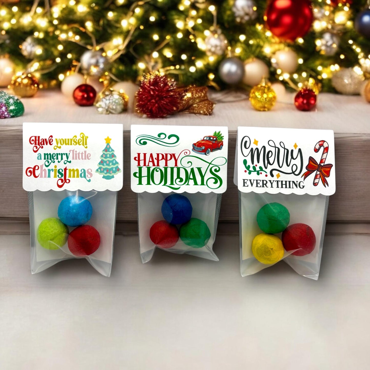 Three Christmas seed bomb 3-packs on a ledge in front of a Christmas tree, with toppers reading 'Have yourself a merry little Christmas', 'HAPPY HOLIDAYS' with a red truck, and 'Merry EVERYTHING' with a candy cane