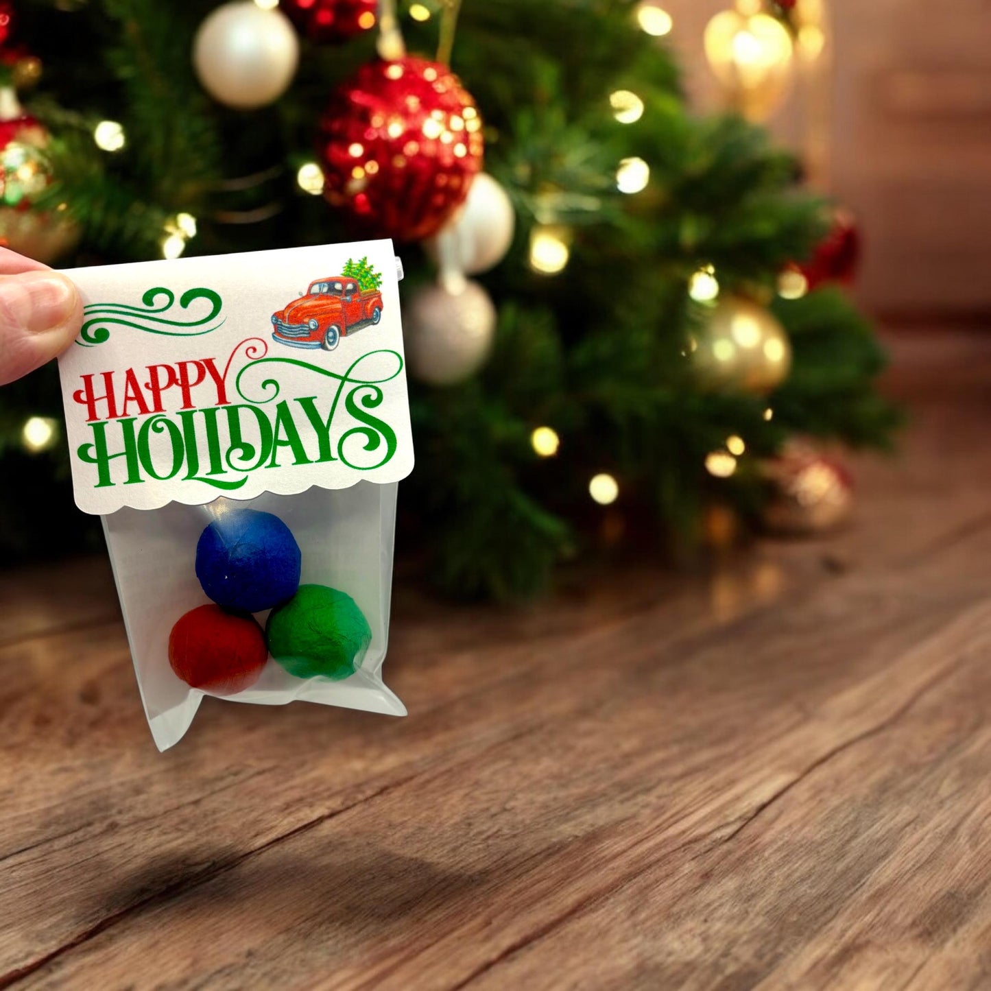 Hand holding a Christmas seed bomb 3-pack with blue, red and green seed bombs and a 'HAPPY HOLIDAYS' topper showing a red truck carrying a Christmas tree, in front of a decorated tree